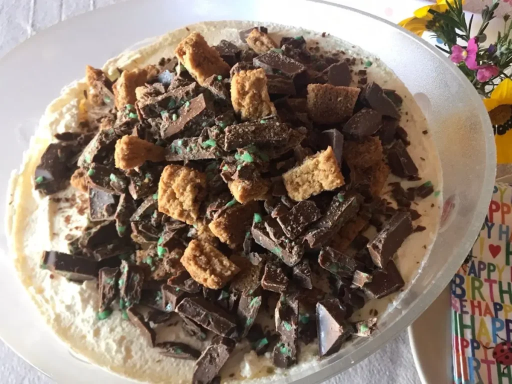 Glass bowl filled with peppermint crisp pudding, topped with chopped mint chocolate and biscuit pieces, set on a beige tablecloth with birthday-themed napkins and spoons.
