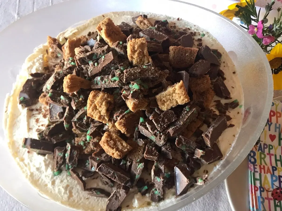 Glass bowl filled with peppermint crisp pudding, topped with chopped mint chocolate and biscuit pieces, set on a beige tablecloth with birthday-themed napkins and spoons.