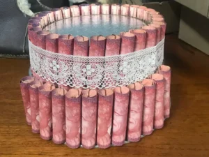 Two-tier decorative money cake made with rolled pink currency notes arranged vertically around each tier, secured with paper clips and a white lace ribbon, and displayed on a wooden surface.