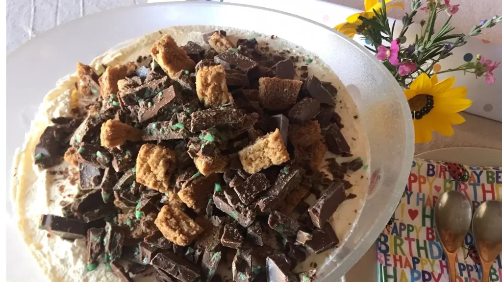 Glass bowl filled with peppermint crisp pudding, topped with chopped mint chocolate and biscuit pieces, set on a beige tablecloth with birthday-themed napkins and spoons.