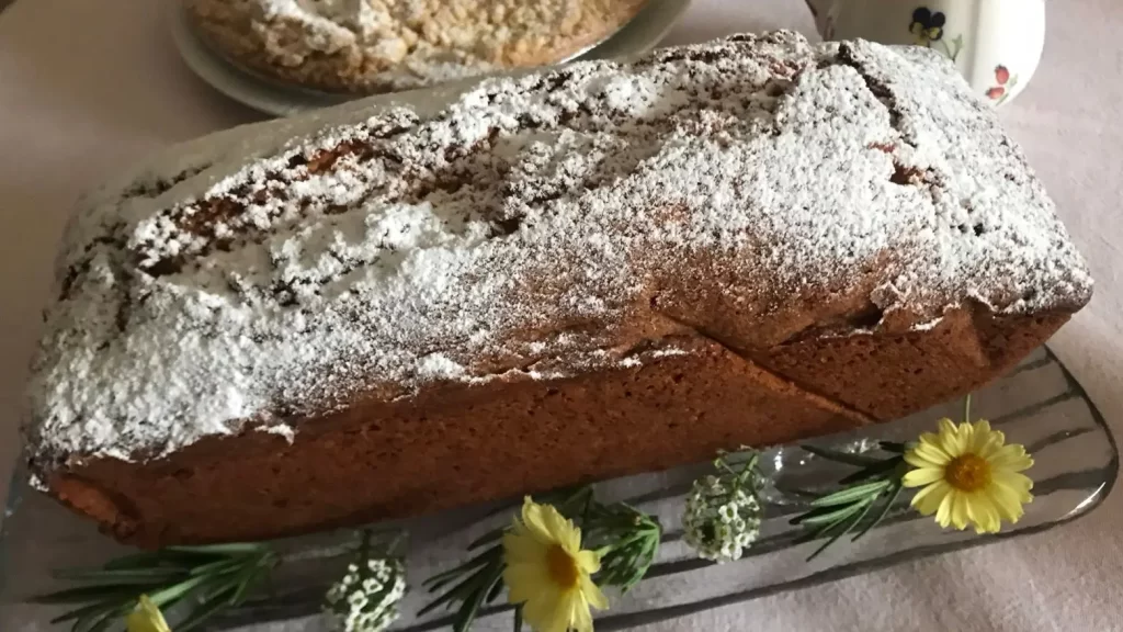 German Ottilienkuchen dusted with powdered sugar on a glass platter, decorated with daisies and rosemary.