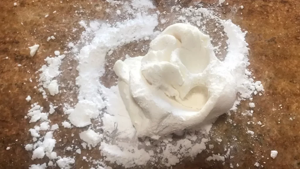 A small mound of homemade white gum paste sits on a lightly dusted surface with powdered sugar scattered around.