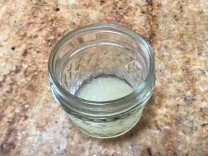 Small glass jar with bloomed gelatine mixture on a speckled kitchen countertop.