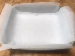 Glass baking dish lined with overhanging parchment paper on a kitchen counter ready to pour in South African fudge mixture.