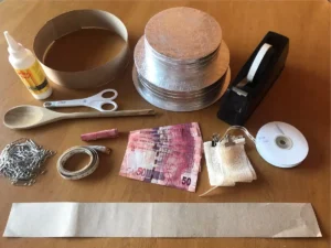 Materials laid out for assembling a two-tiered money cake, including cake boards, cardboard strips, glue, tape, paper clips, ribbon, R50 South African banknotes, and crafting tools.