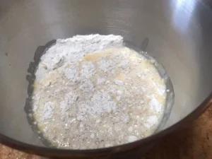Ingredients for cinnamon roll dough in a mixing bowl, including flour, sugar yeast, and melted butter and mlik.