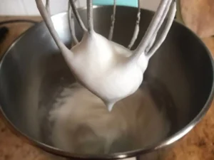 Stiffly whipped egg whites forming peaks on a metal whisk over a mixing bowl for the Strawberries and Cream Swiss Roll Recipe.