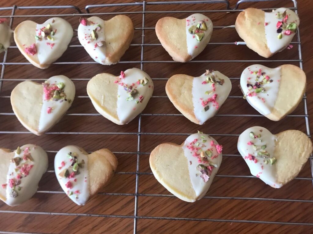 Heart-shaped shortbread cookies half-dipped in white chocolate, decorated with chopped pistachios and crushed freeze-dried strawberries, arranged on a cooling rack.