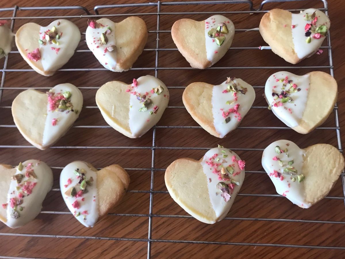 Heart-shaped shortbread cookies half-dipped in white chocolate, decorated with chopped pistachios and crushed freeze-dried strawberries, arranged on a cooling rack.