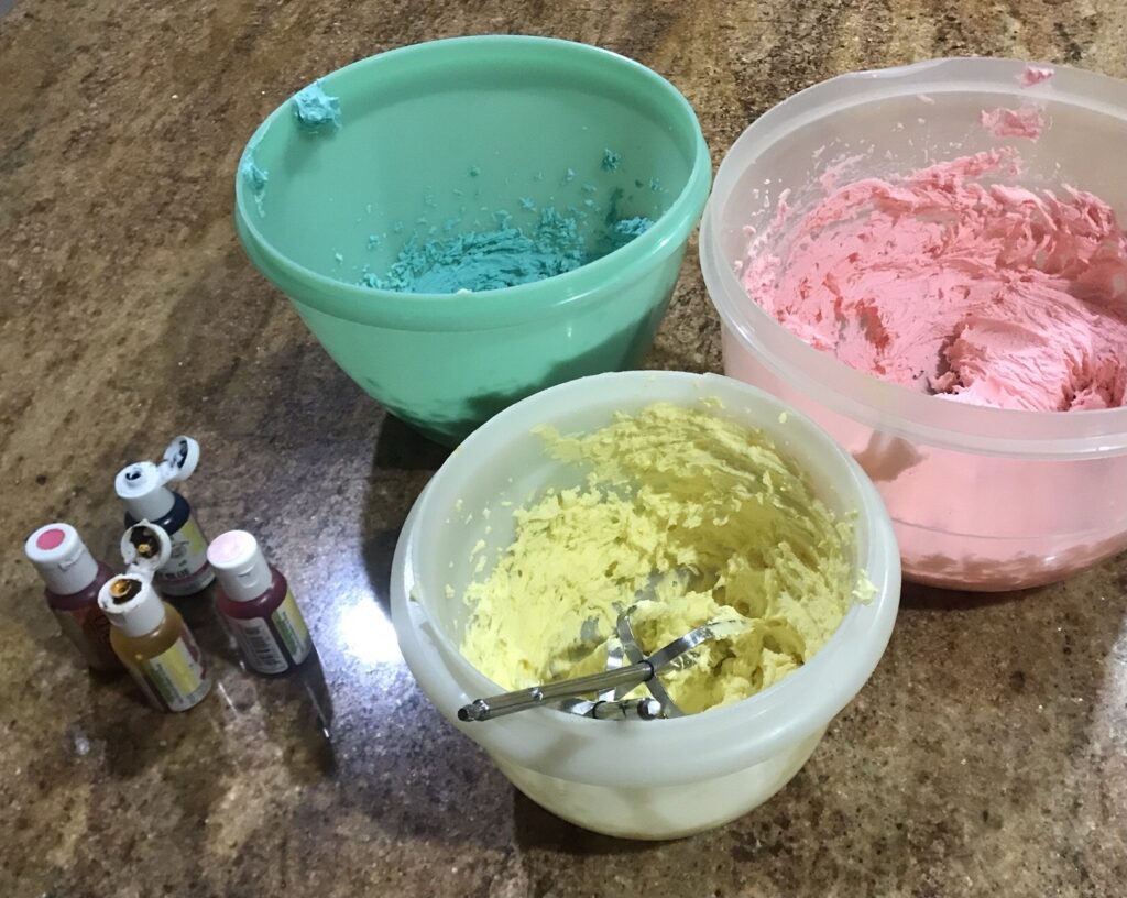 Three bowls of coloured butter icing in pink, yellow, and blue on a kitchen counter, with gel food colour bottles nearby.
