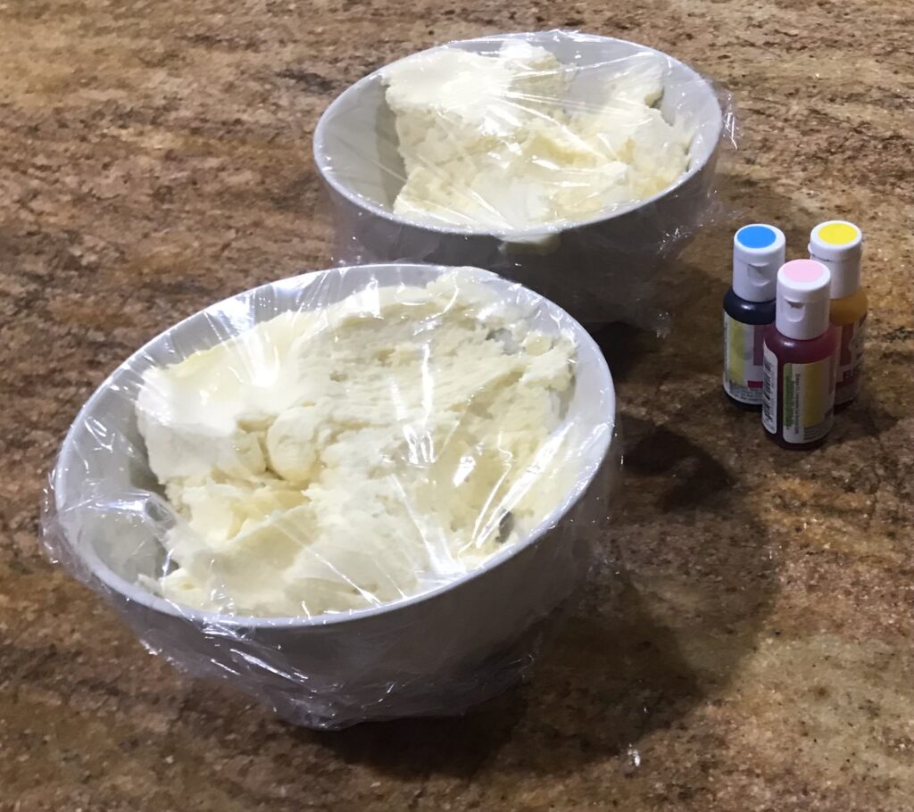 Two bowls of freshly made vanilla butter icing covered in plastic wrap, with gel food colouring bottles beside them.