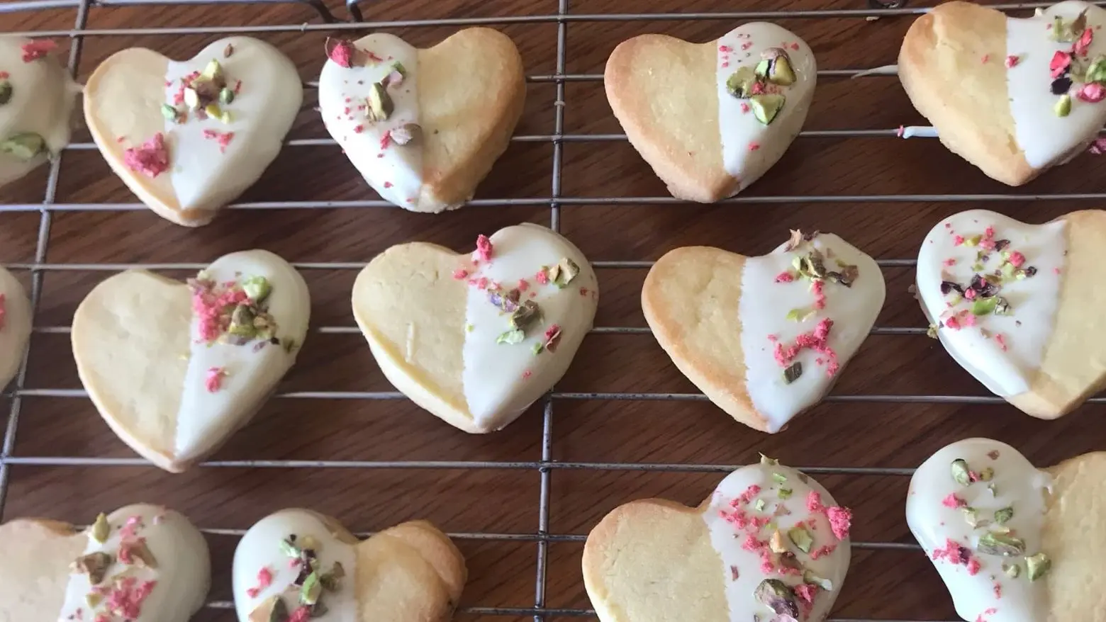 Heart-shaped shortbread cookies half-dipped in white chocolate, decorated with chopped pistachios and crushed freeze-dried strawberries, arranged on a cooling rack.