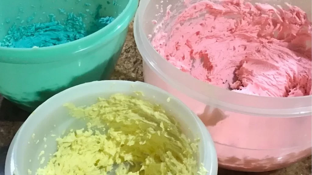 Three bowls of coloured buttercream in pink, yellow, and blue on a kitchen counter, with gel food colour bottles nearby.