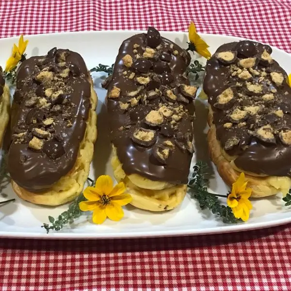 Homemade chocolate éclairs filled with vanilla pastry cream, topped with glossy chocolate and crushed Chuckles/Maltesers served on a white platter with yellow flowers.