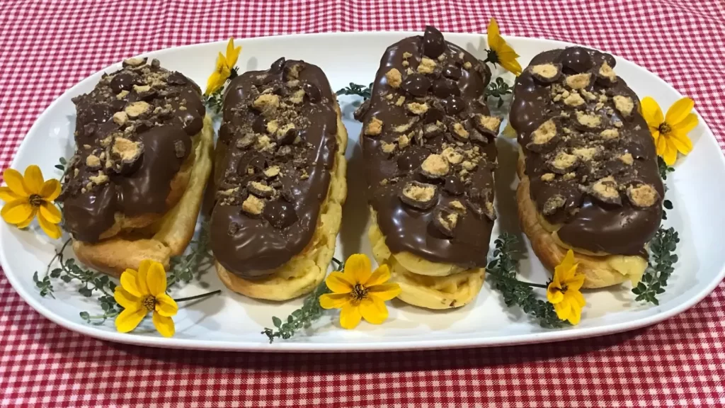 Homemade chocolate éclairs filled with vanilla pastry cream, topped with glossy chocolate and crushed Chuckles/Maltesers served on a white platter with yellow flowers.