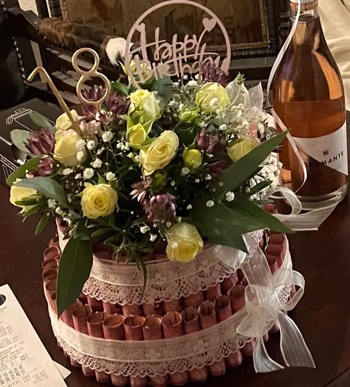 Two-tier money cake with lace ribbon, yellow roses, purple flowers, and a Happy Birthday topper—for milestone celebrations.