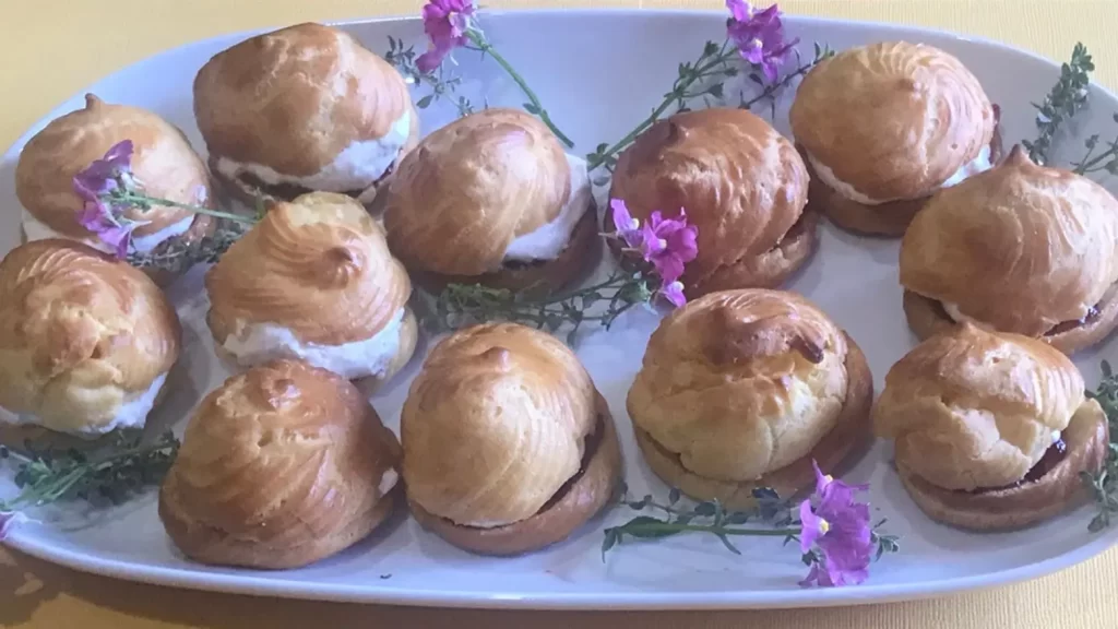 Golden-brown cream puffs (Windbeutel) filled with whipped cream and strawberry jam on a white platter, decorated with purple flowers — classic choux pastry recipe.