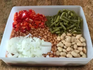 Chopped red pepper, onion, baked beans, green beans, and butter beans arranged separately in a rectangular container before mixing for a three bean salad.