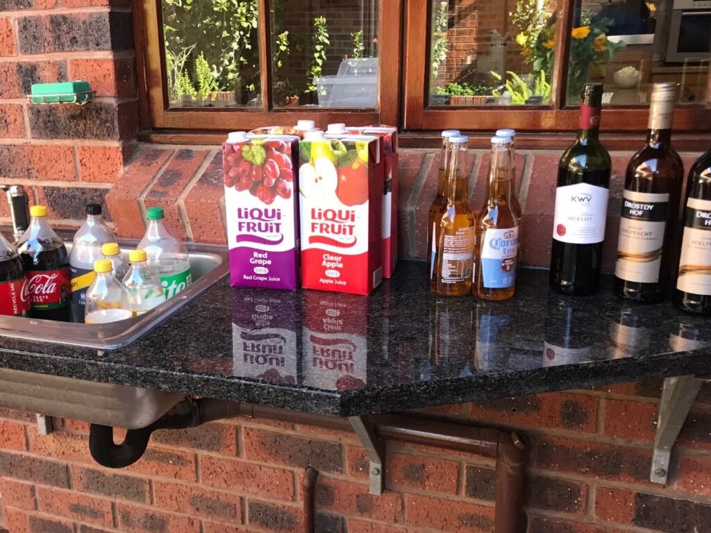 Outdoor bar area with assorted wines, beers, soft drinks, and fruit juices arranged on a black granite counter.