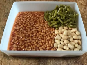 Baked beans, green beans, and butter beans arranged separately in a rectangular container before mixing for a three bean salad.