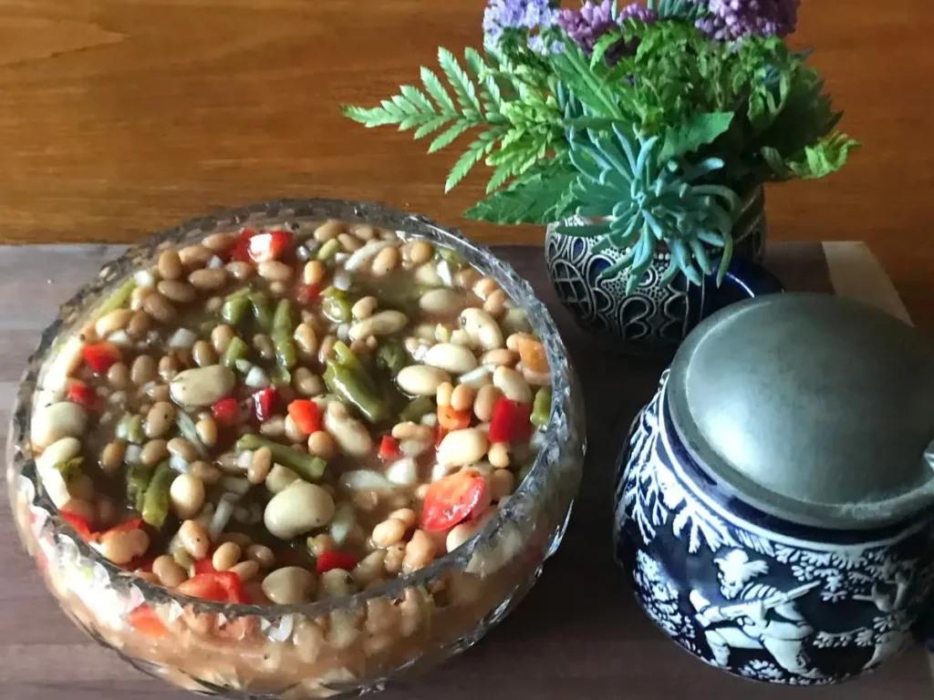 Three bean salad in a glass bowl with baked beans, green beans, butter beans, red pepper, and onion, served on a wooden board beside a blue patterned teapot and a small vase with greenery.