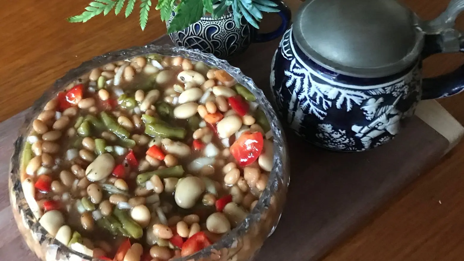 Three bean salad in a glass bowl with baked beans, green beans, butter beans, red pepper, and onion, served on a wooden board beside a blue patterned beer mug and a small vase with greenery.