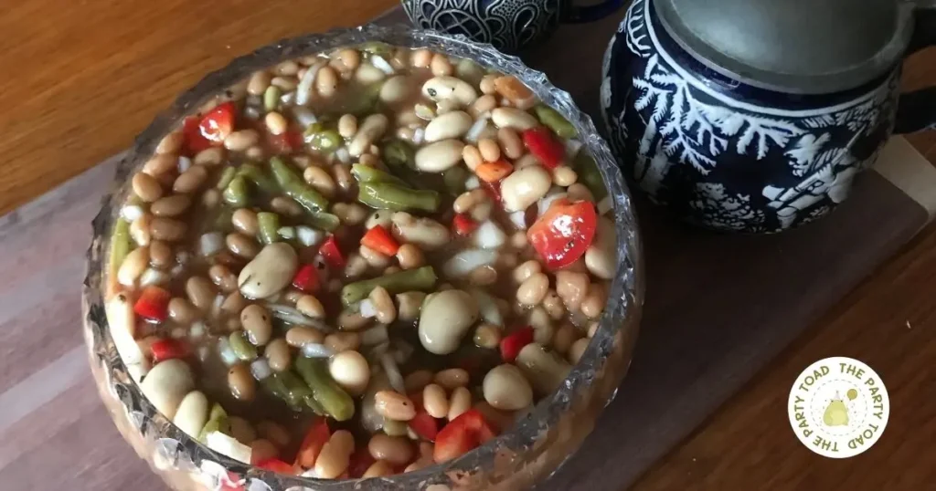 Three bean salad in a glass bowl with baked beans, green beans, butter beans, red pepper, and onion, served on a wooden board beside a blue patterned beer mug and a small vase with greenery.