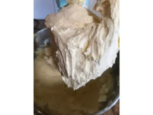 Close-up of medium consistency buttercream on a mixer paddle with a bowl of icing underneath.