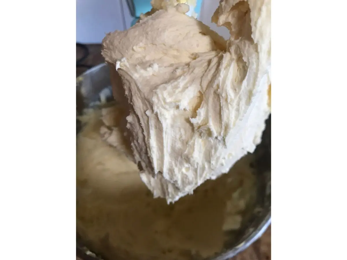 Close-up of medium consistency buttercream on a mixer paddle with a bowl of icing underneath.