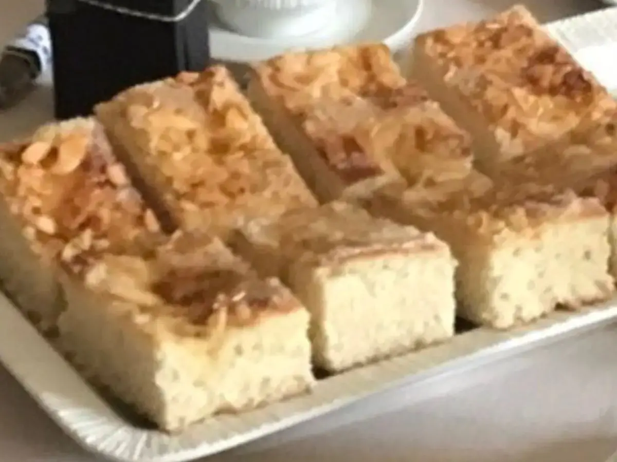 Slices of Butterkuchen, a traditional German sheet cake, with a golden crust, crunchy almond topping, and a soft, fluffy interior, served on a platter with a cup of coffee in the background.