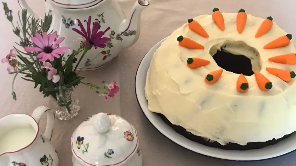 Ring-shaped carrot cake topped with cream cheese frosting and decorated with mini carrots, with floral china tea set and a small vase of garden flowers.