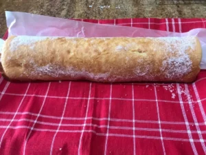 Freshly baked Swiss roll dusted with sugar, resting on parchment paper and a red checkered tea towel.