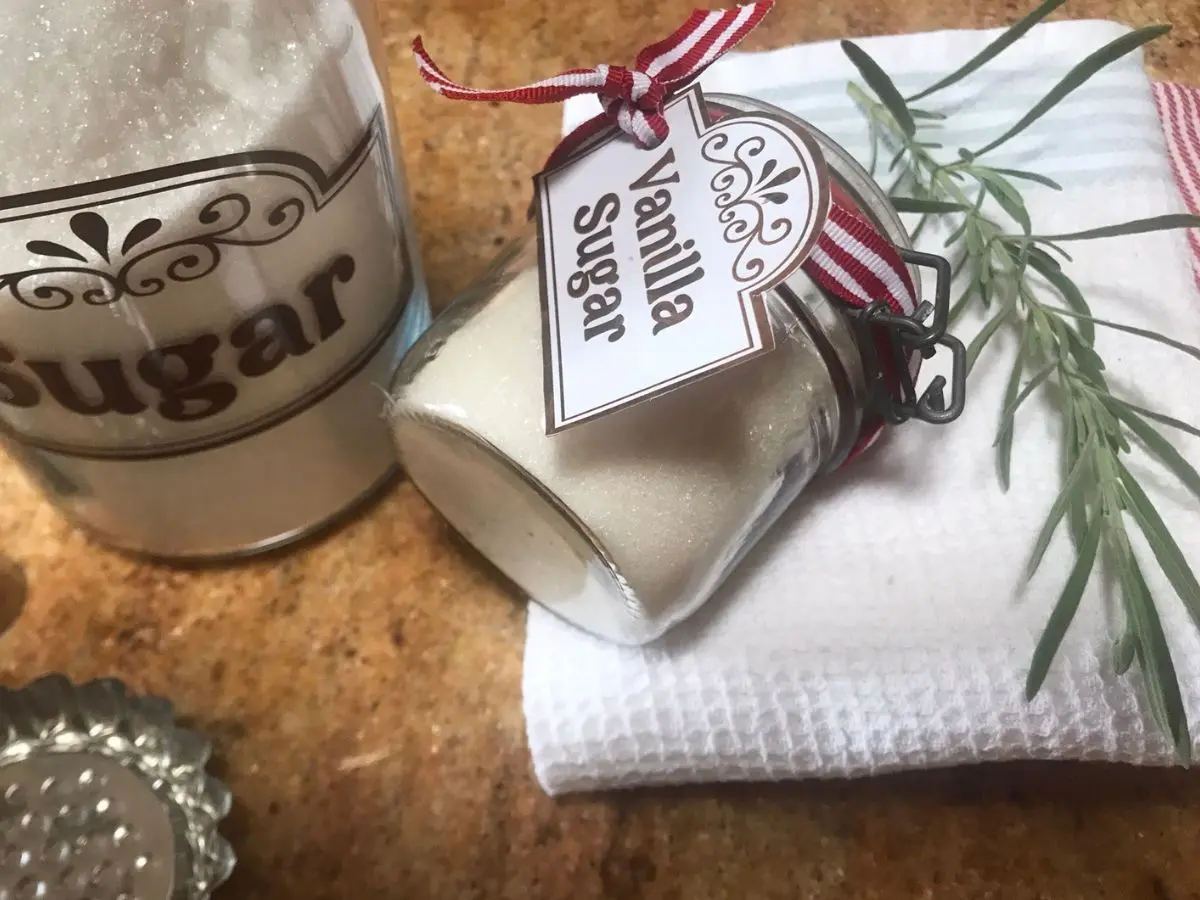 A glass jar of homemade vanilla sugar with a decorative label and red ribbon, placed on a white towel next to a sprig of thyme, with a larger sugar container in the background.